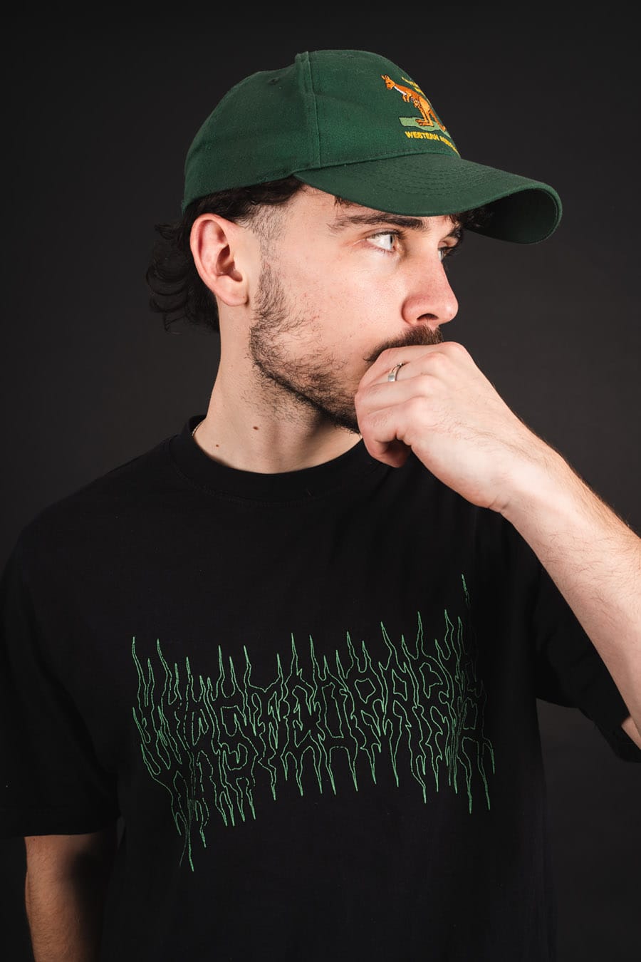 Primate in a green cap and black t-shirt, hand at his chin, against a dark backdrop.