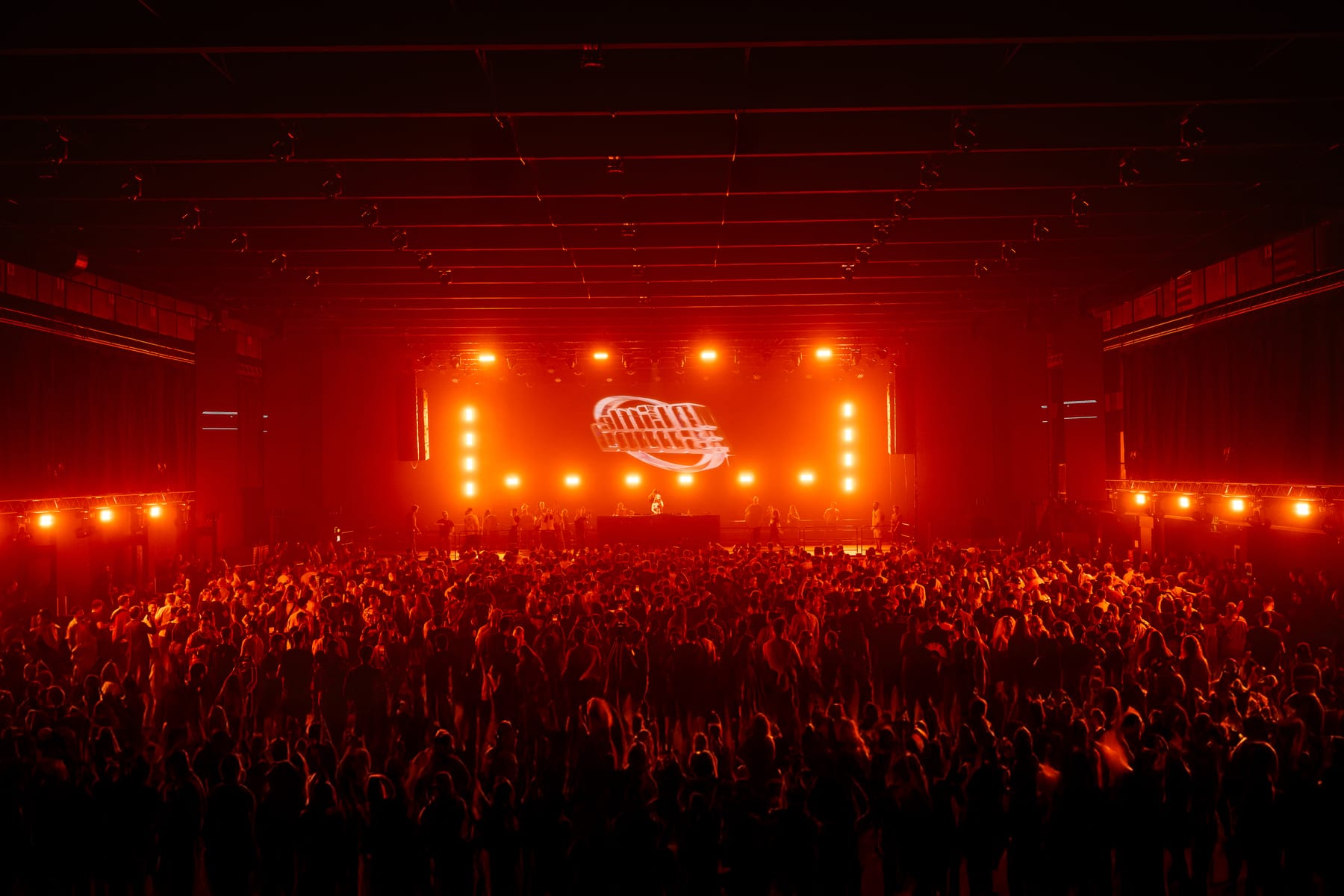 A drum and bass crowd filling a large hall under deep red stage light.